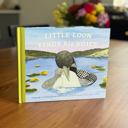 Children's book titled 'Little Loon Finds his Voice' on a wooden surface with flowers in the background.