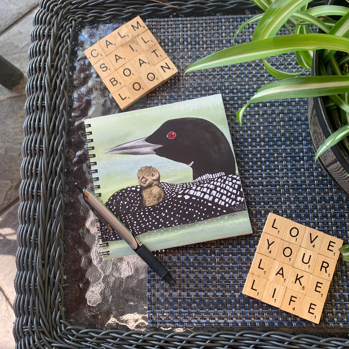 A top-down view of a dark woven wicker tray on a stone patio, styled with a spiral notebook featuring a loon illustration, Scrabble tiles, a pen, and a potted spider plant.